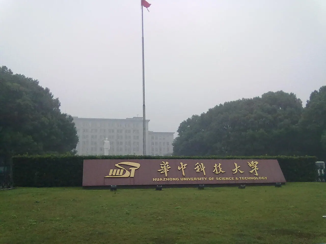 Biển tên Đại học Khoa học và Công nghệ Hoa Trung trước thảm cỏ, hàng cây và tòa nhà phía sau.